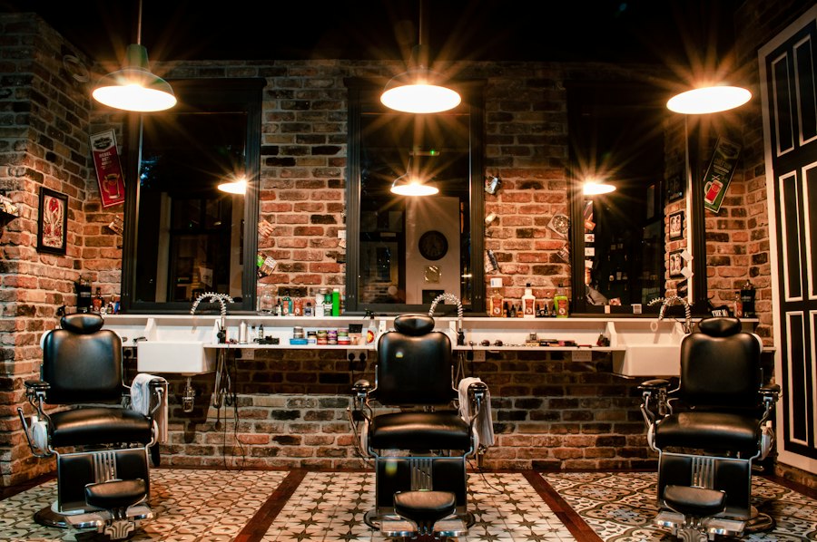 Three black leather barber chairs under warm pendant lights against brick walls